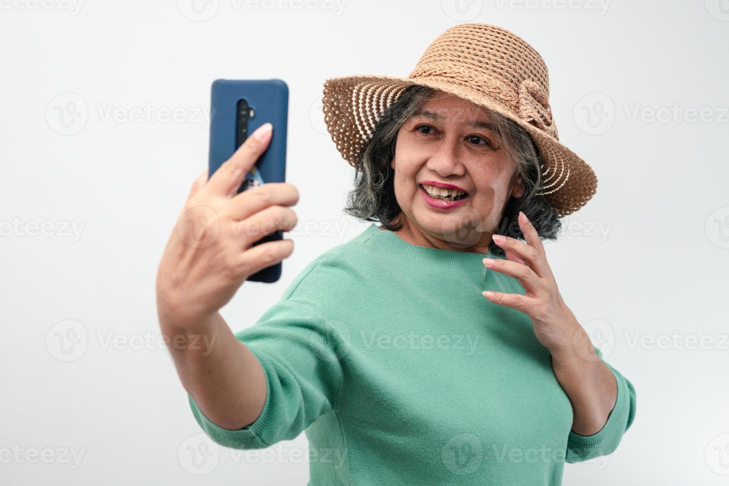 Portrait of happy Asian senior woman wearing sunglasses, a hat, and holding smartphone for a selfie, standing isolated on white background. Concept of tourist enjoy and fun valentine day after retire photo