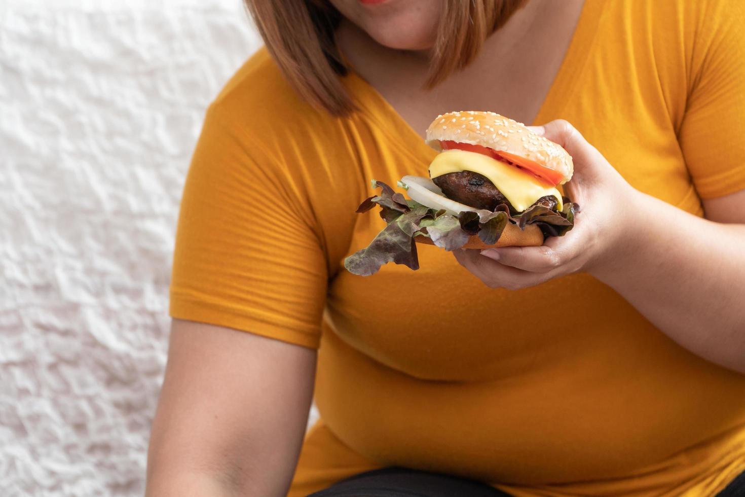 Hungry overweight woman smiling and holding hamburger and sitting in the bedroom, her very happy and enjoy to eat fast food. Concept of binge eating disorder BED. photo