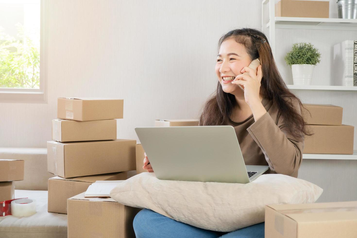 Happy Asian young entrepreneur, talking on the smartphone with customer for tack order, She smile for sales success at home office, Concept of merchant business online and eCommerce photo