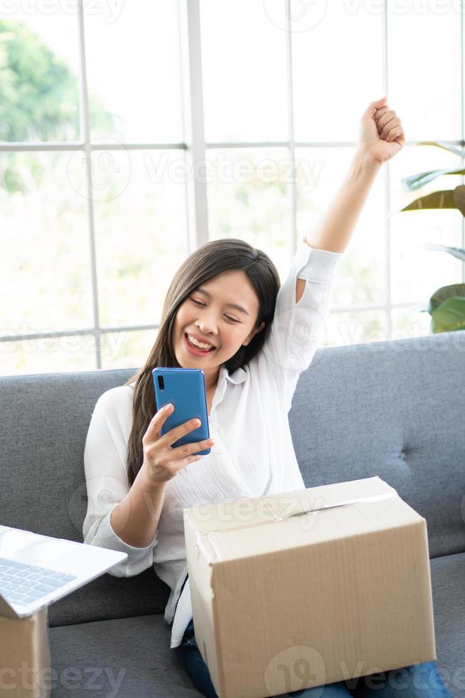 Happy young Asian woman entrepreneur, Smile for sales success after checking order from online shopping store in a smartphone at home office, Concept of merchant business online and eCommerce photo