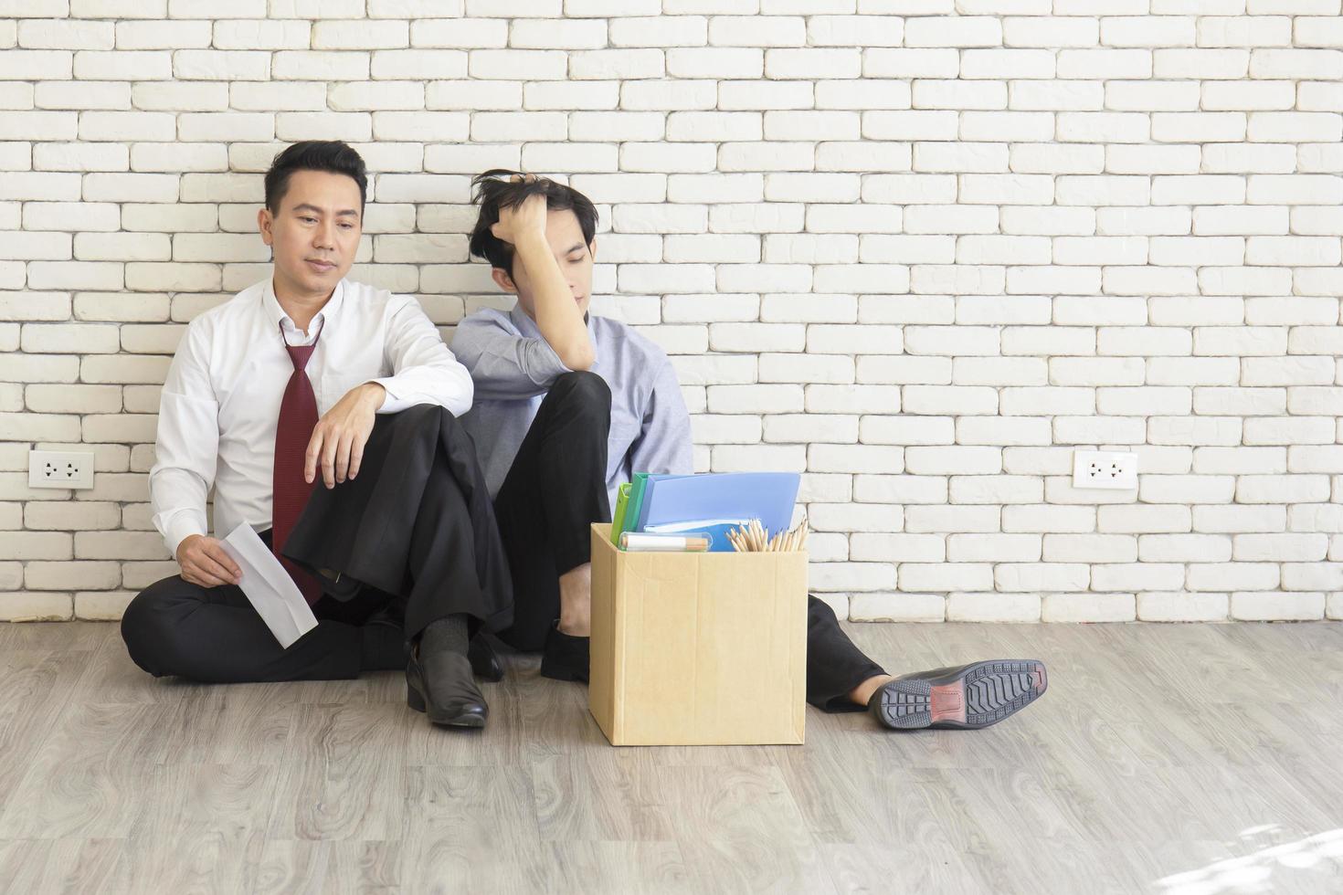 Two male employees sat on the floor talking in front of each other with a cardboard box for personal use after being fired from work. photo
