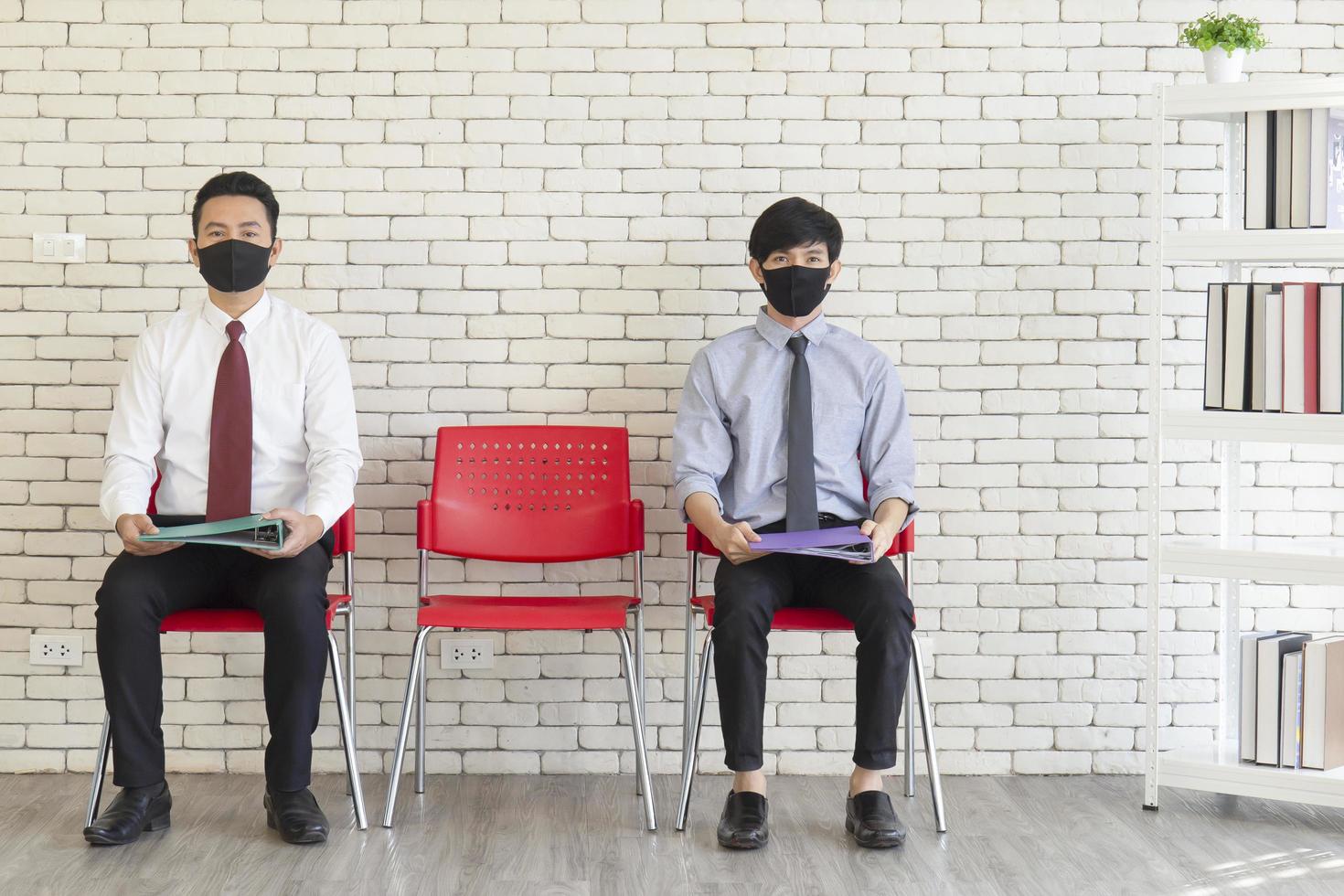 Two Asian men sit in red plastic chairs spaced out with Matt to cover their mouths to prevent COVID-19 waiting for a job interview. photo