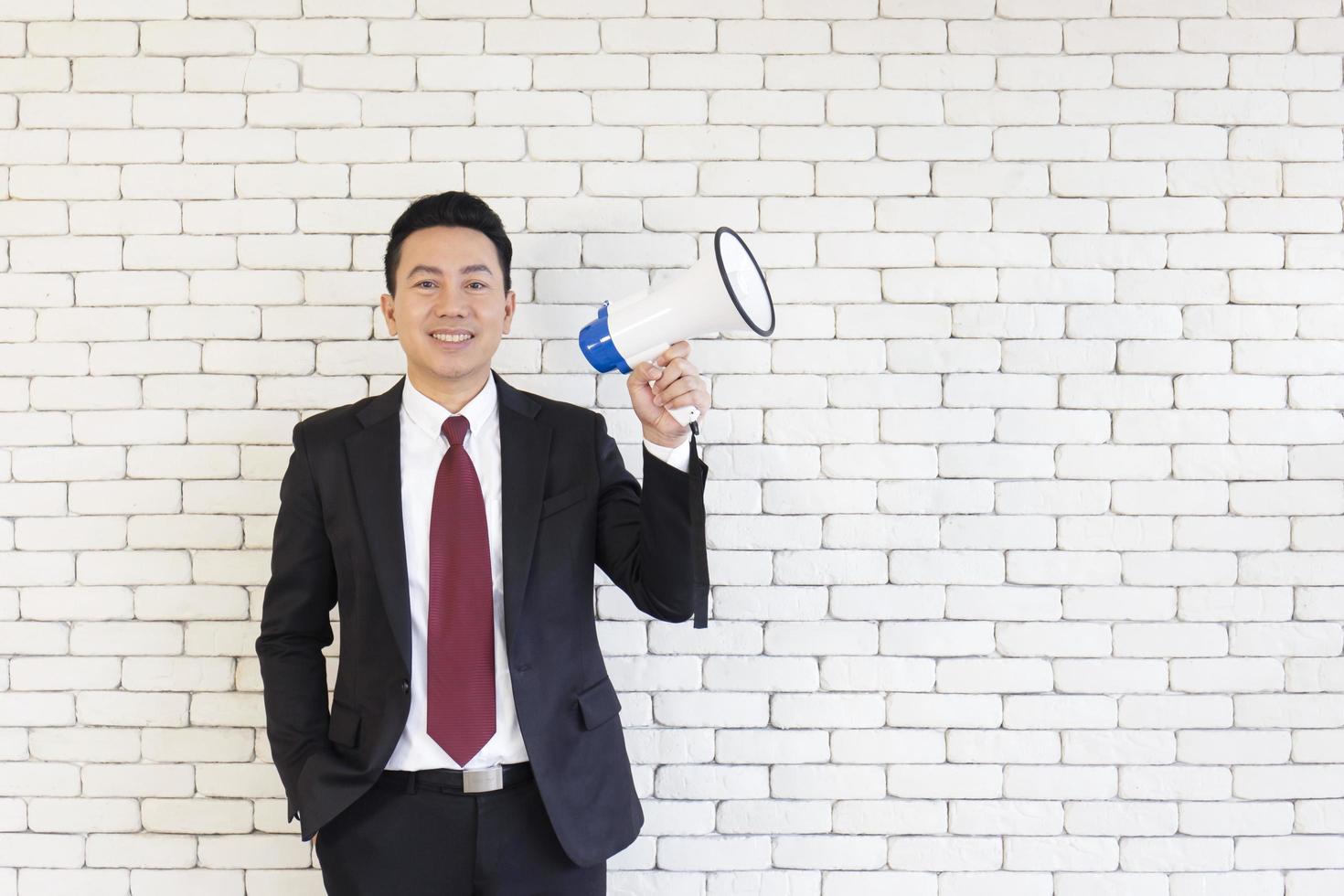 An Asian man in a suit holds a megaphone on a white brick wall. photo