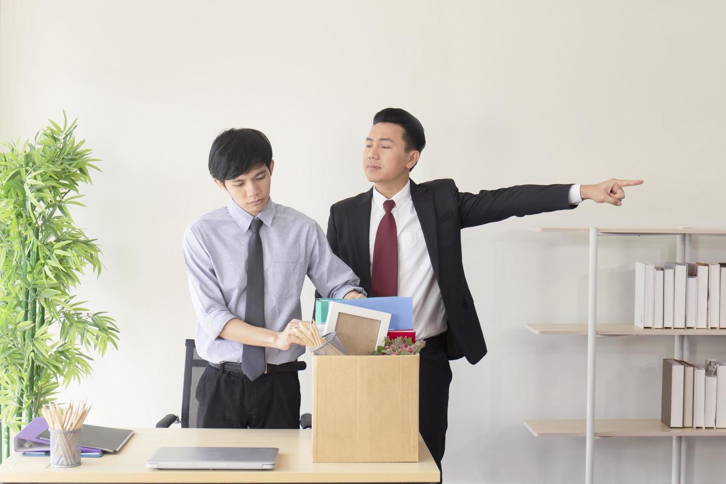 An Asian employee was fired from his job and was packing his personal belongings in paper boxes with a manager standing by him. photo