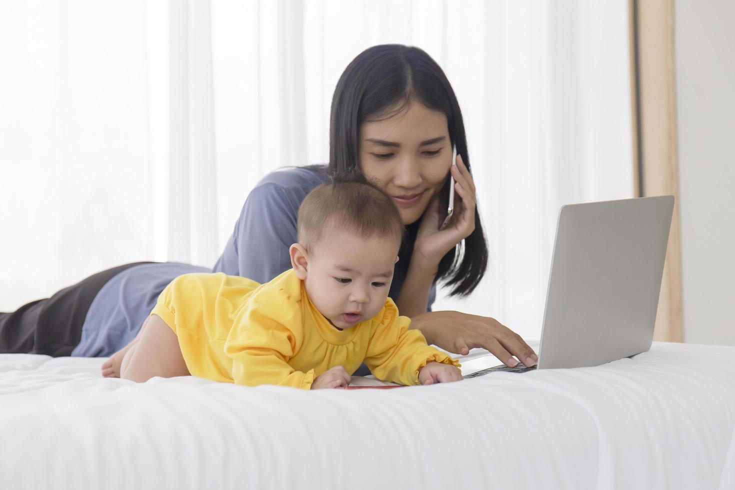 An Asian baby is playing next to her mother in the bed, with her mother talking on the cell phone. photo