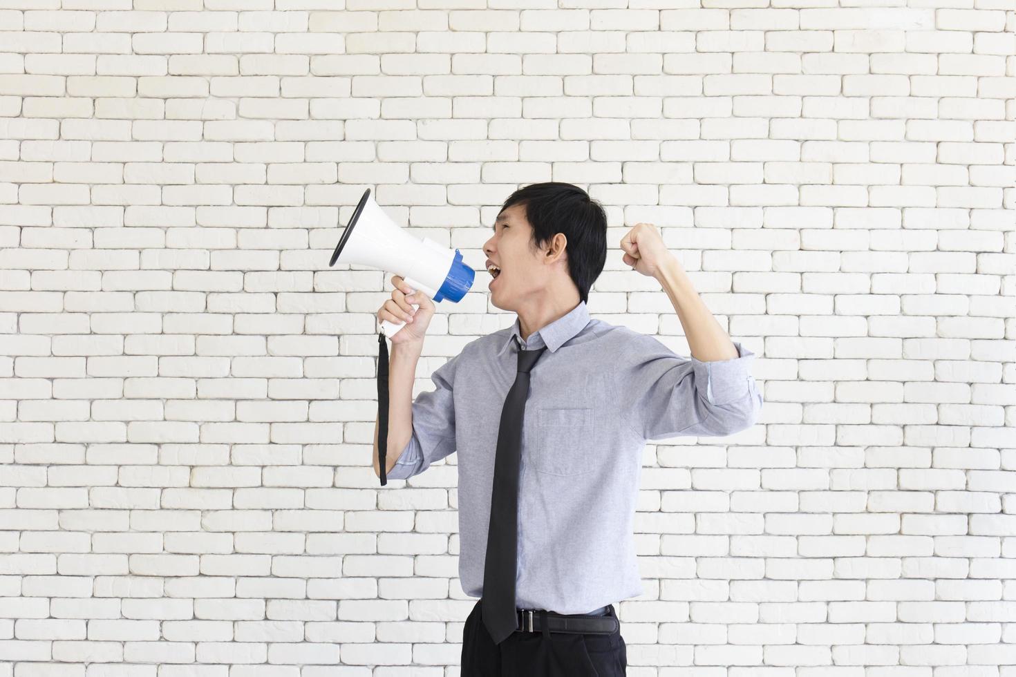 An Asian man stands holding a blue and white megaphone behind a white brick wall. photo