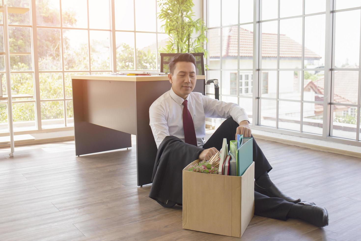 An Asian male company worker sits on the floor beside a desk with a cardboard box containing his personal belongings. photo