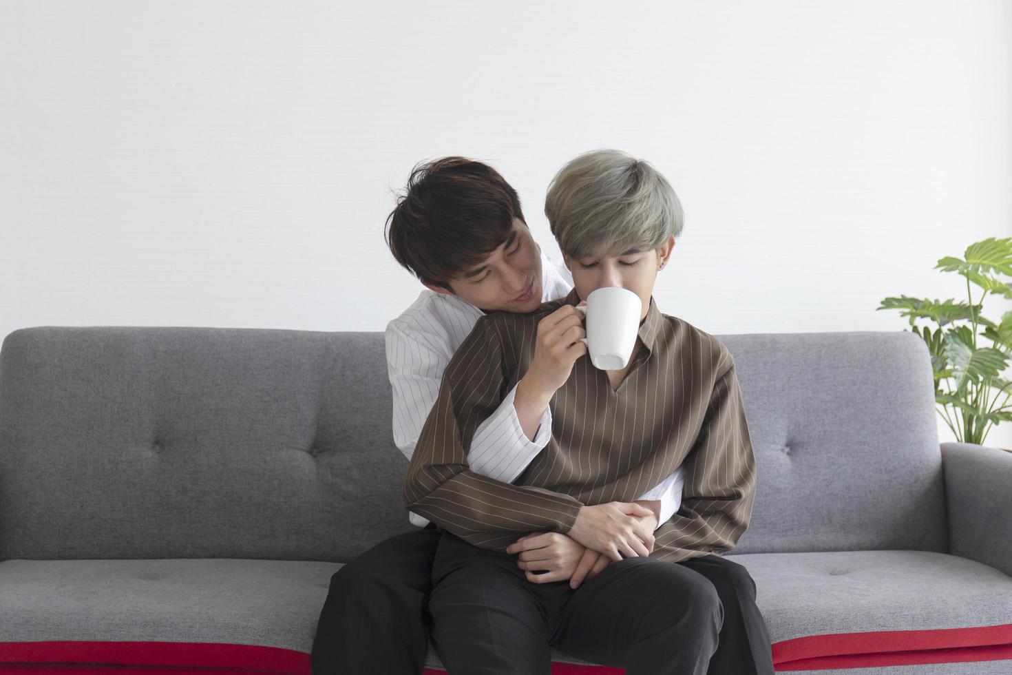 A male couple with an Asian man cuddling on the sofa and feeding coffee. photo