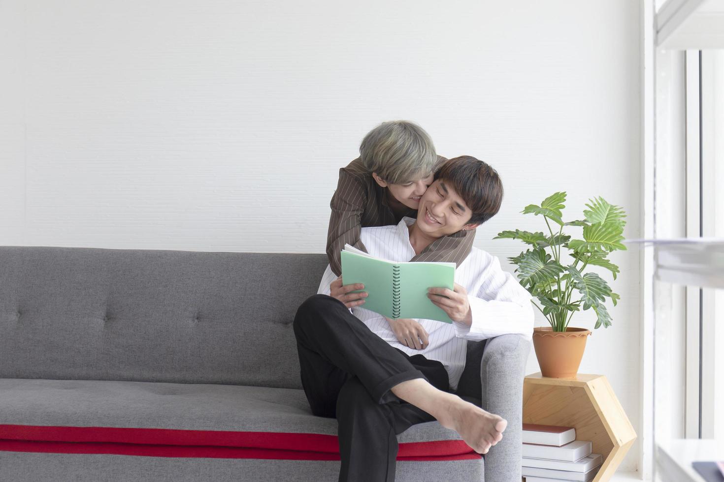 una pareja masculina con un hombre asiático acurrucado en el sofá con su amor mutuo y el otro leyendo un libro. foto