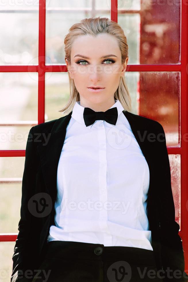 retrato de una hermosa y elegante mujer rubia con traje negro de hombre cerca de una cabina telefónica roja foto