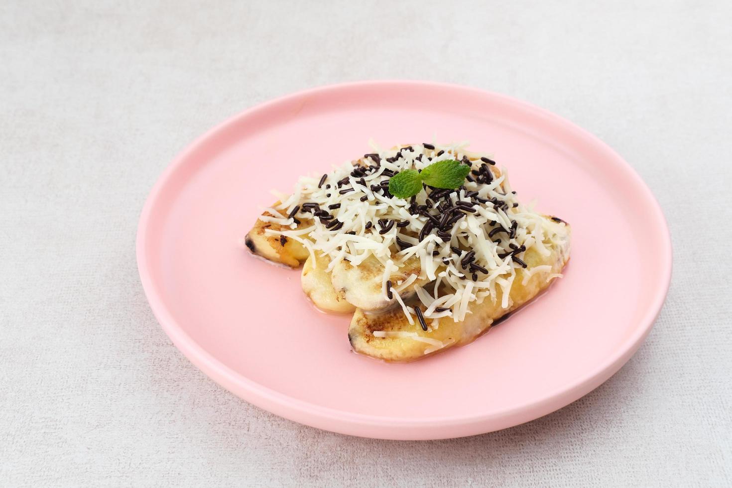 pisang panggang, plátano a la parrilla cubierto con chispas de chocolate, queso rallado y dulce de leche blanca. servido en plato con hojas de menta sobre fondo gris. foto