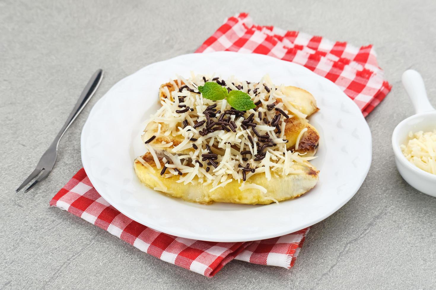 Pisang panggang, Grilled banana topped with chocolate sprinkles, grated cheese, and sweet white milk. Served on plate with mint leaves on grey background. photo