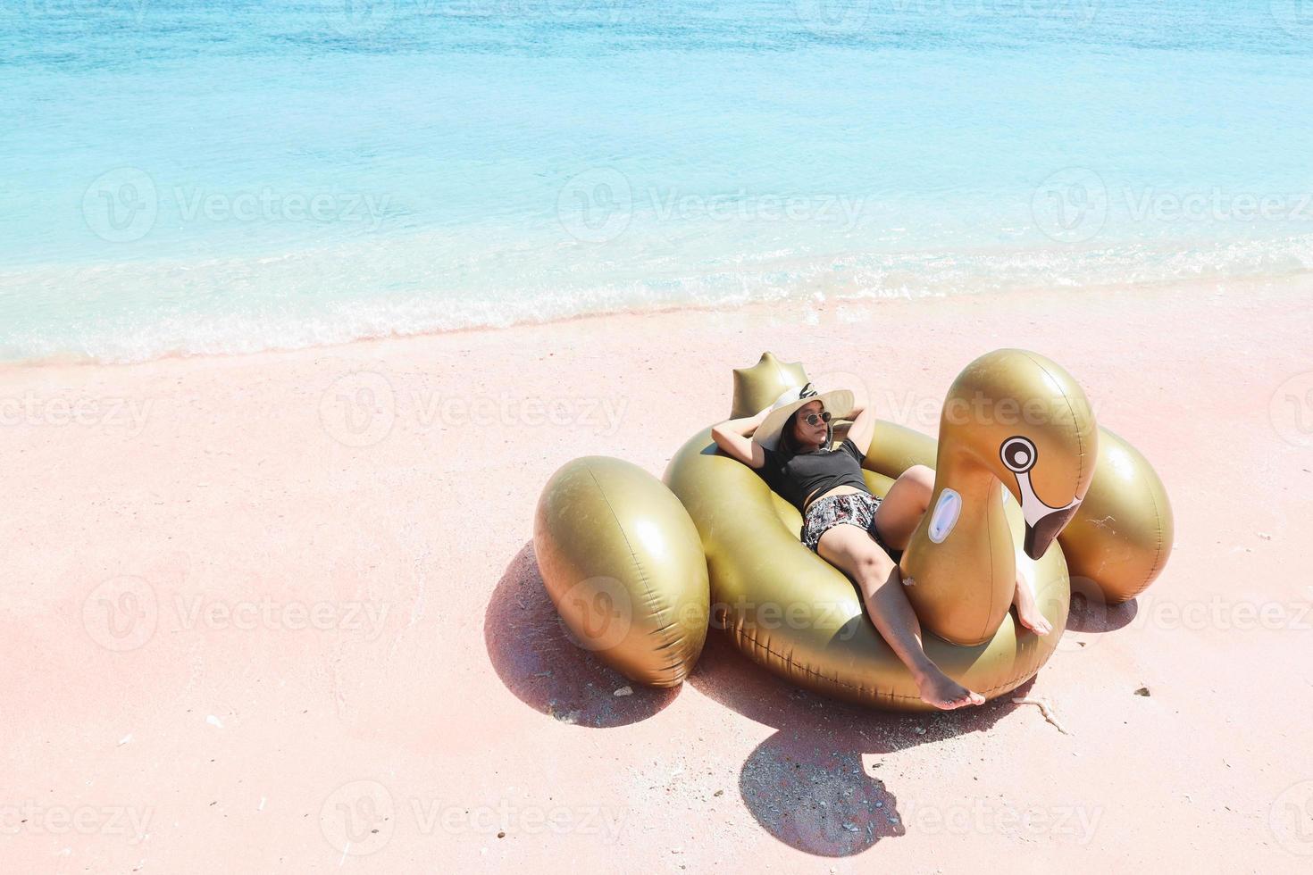 Asian woman in summer hat and sunglasses laying on inflatable swan in pink sandy beach photo