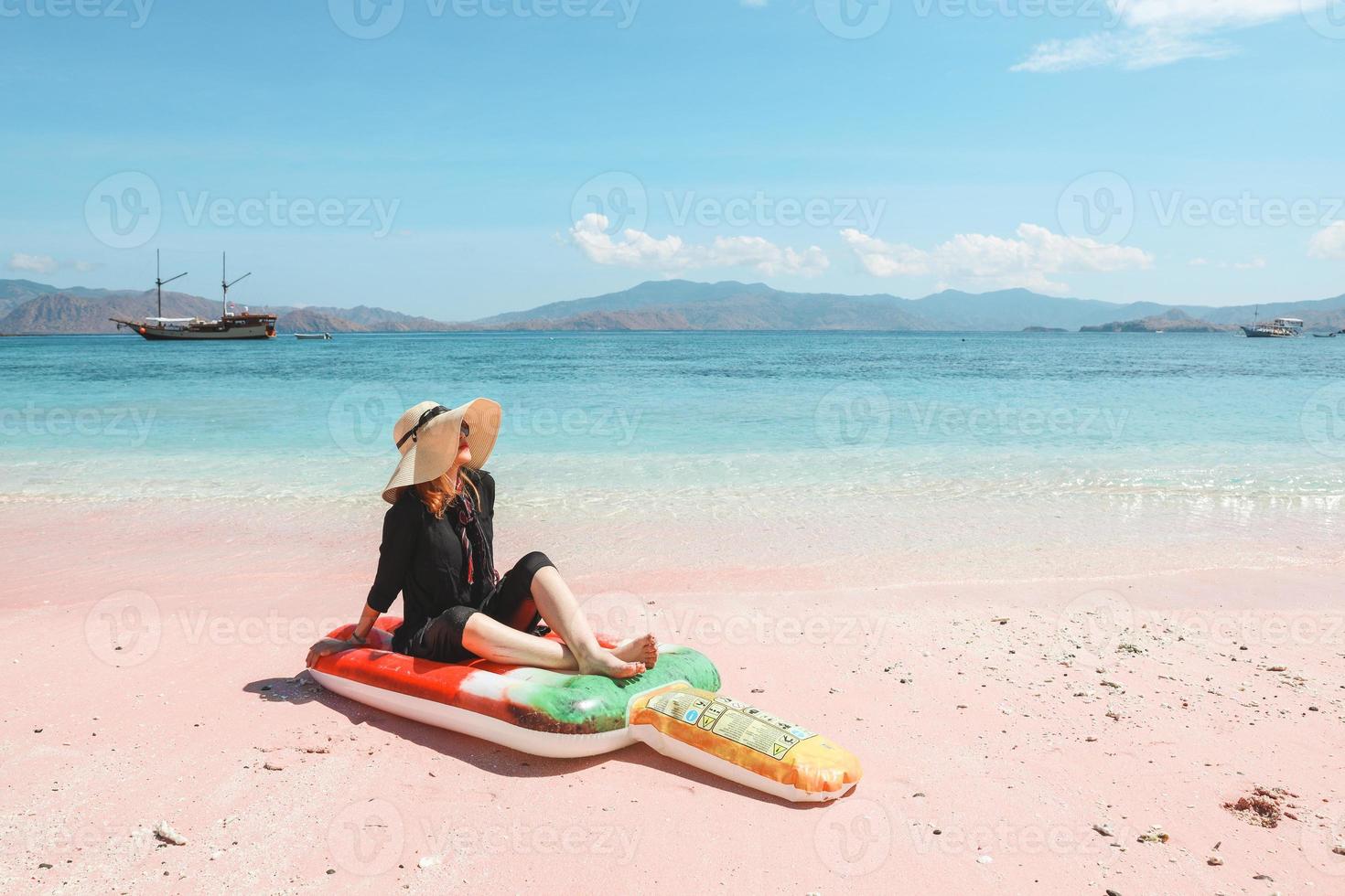 Blonde woman in summer hat and sunglasses relaxing on inflatable mattress in pink sandy beach photo