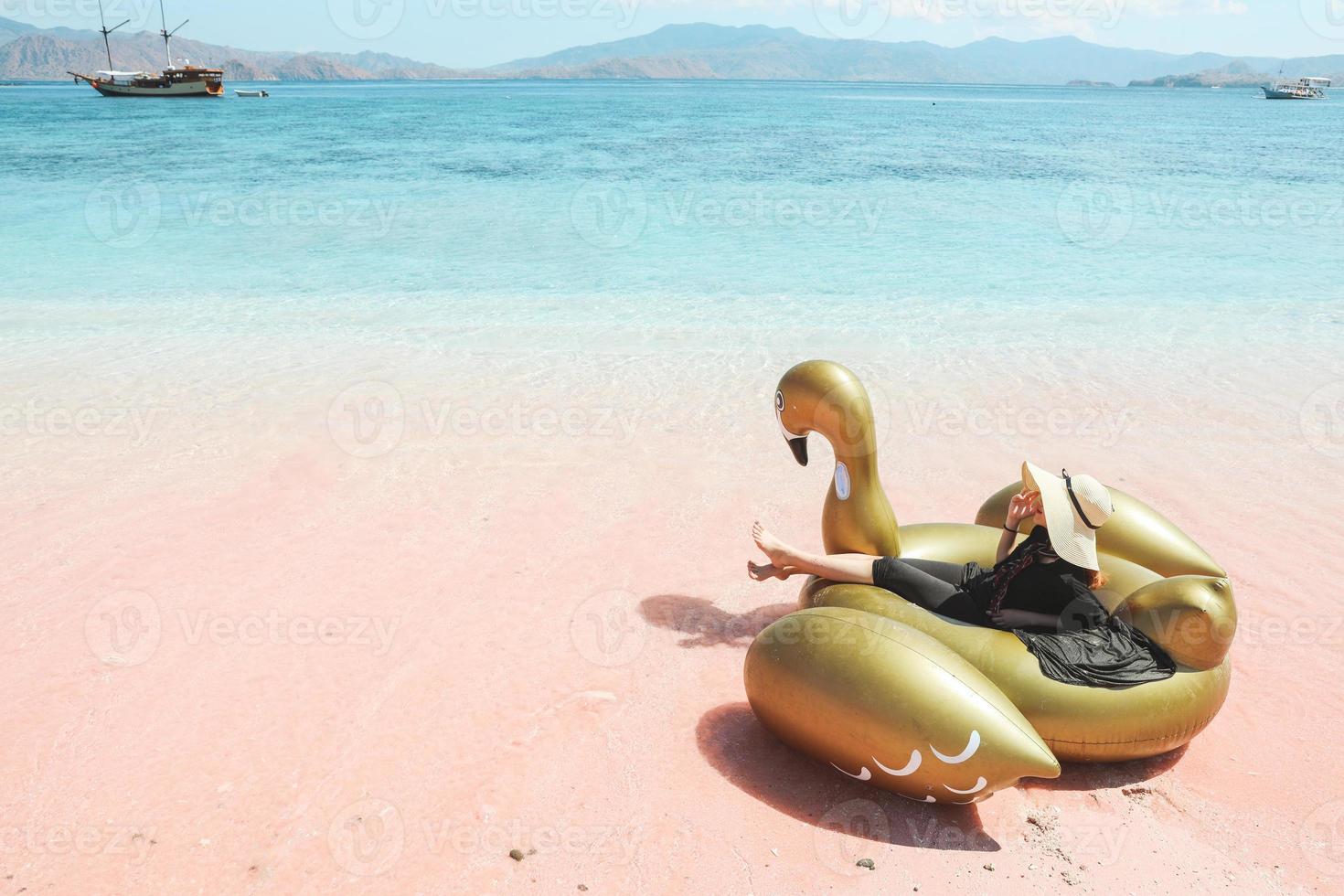 Female tourist in summer hat relaxing on golden inflatable swan on pink sandy beach photo