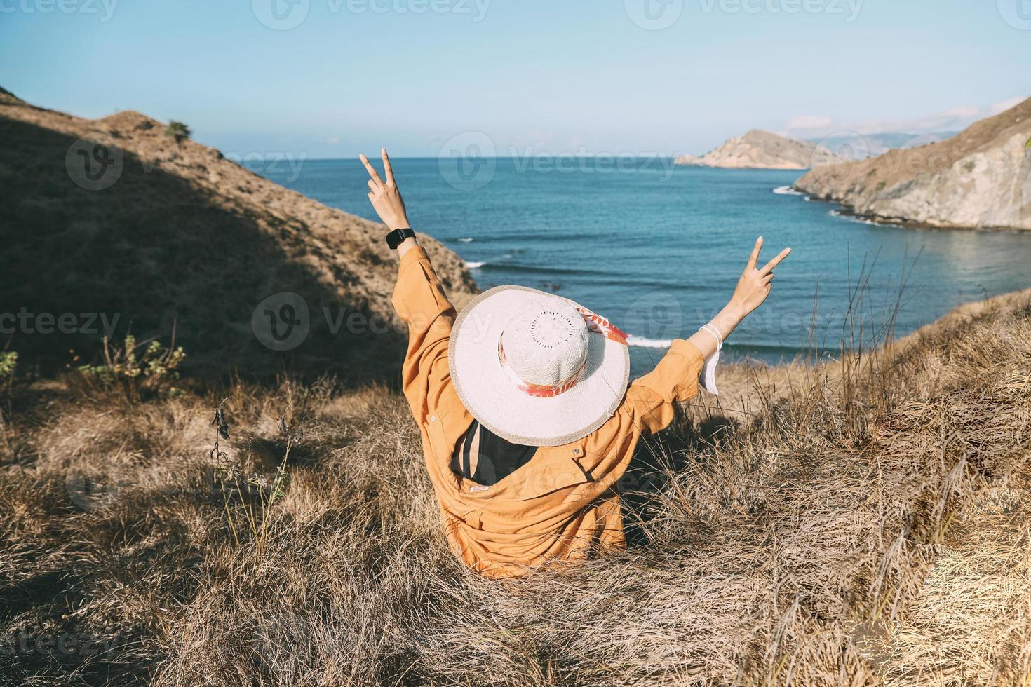 Happy female tourist wearing a summer hat sitting and raising her hands symbolizing peace with a beautiful view photo