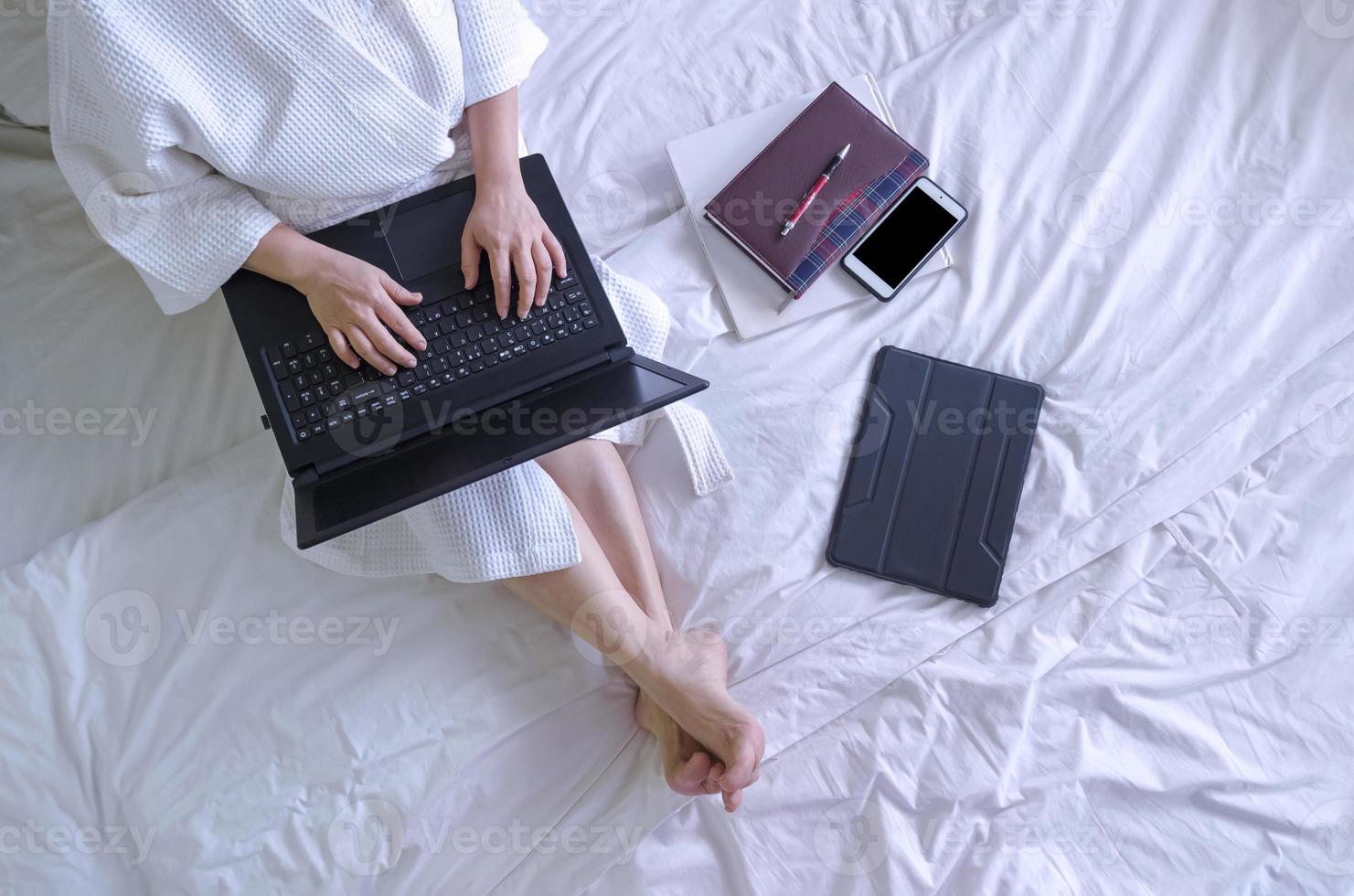 Top view of woman in white bathrobe using laptop computer with book and various device on bed in her bedroom photo