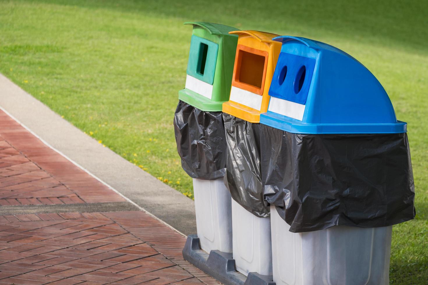 3 cubos de basura de reciclaje coloridos para la clasificación separada de residuos en el pavimento de baldosas de piedra con césped verde en el área del parque público foto