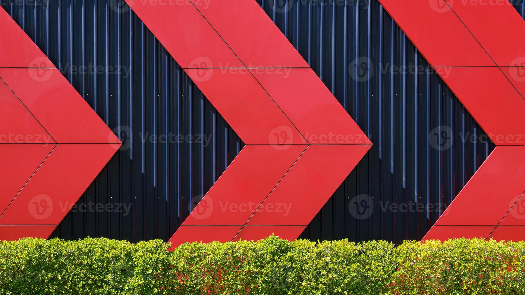 Front view of red arrows pattern on black corrugated metal wall behind green bush fence in exterior architecture decorations design concept photo