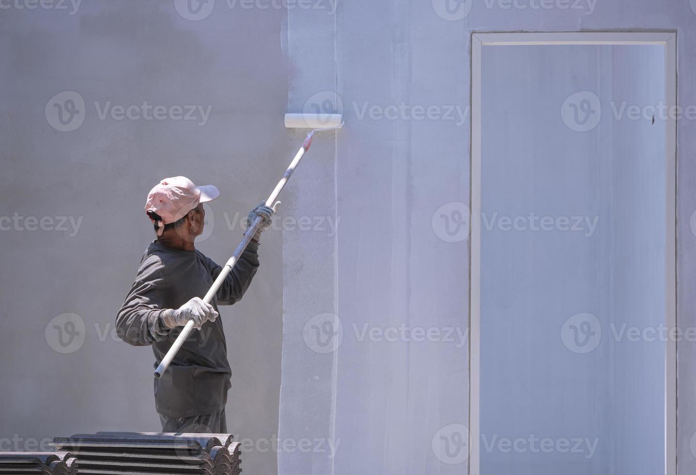 trabajador constructor asiático que usa un cepillo de rodillos de mango largo para pintar el color blanco de la imprimación en la pared de hormigón dentro del sitio de construcción de la casa foto