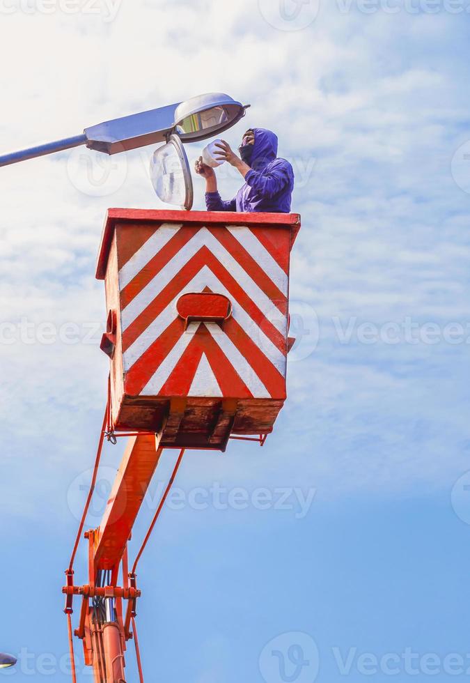 Low angle view of Asian electrician in bucket boom truck is repairing street lamppost against blue sky in vertical frame photo