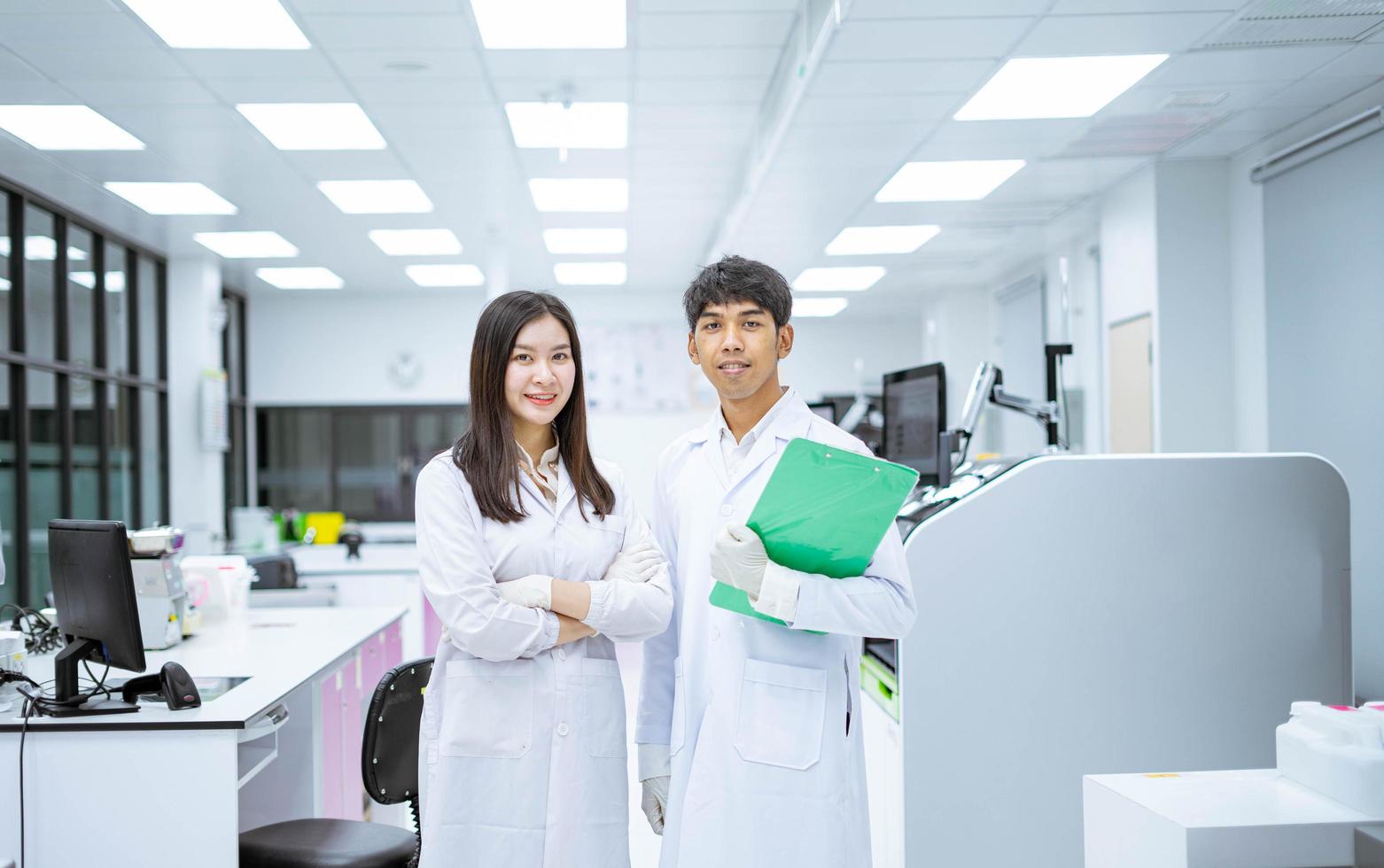 two young smiling scientist in white lab coat standing with automation blood analyzer at medical laboratory photo