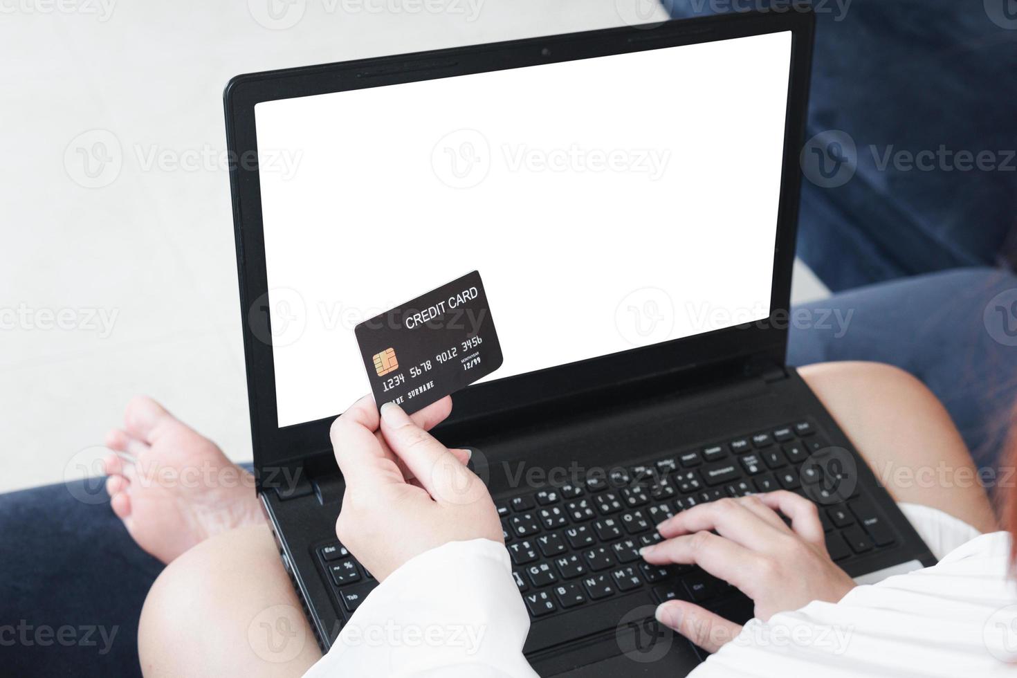 una mujer que tiene una tarjeta de crédito, que paga en línea, en una computadora portátil, sentada en el sofá de la sala de estar, en casa con un camino de recorte. el concepto de compras en línea. cuaderno de pantalla blanca para maqueta foto