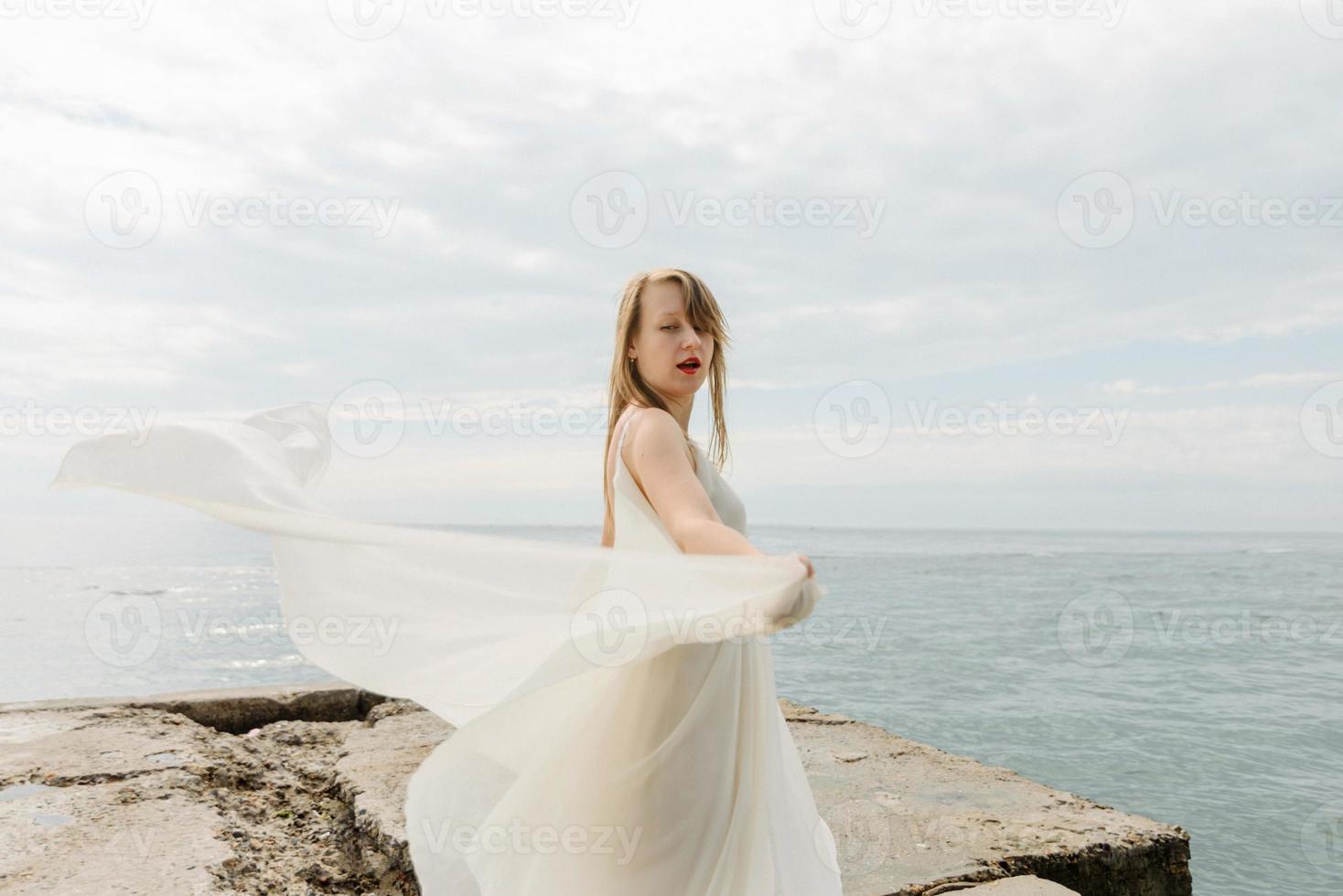 una joven hermosa con un largo vestido color leche camina por la playa y el muelle contra el fondo del mar. foto