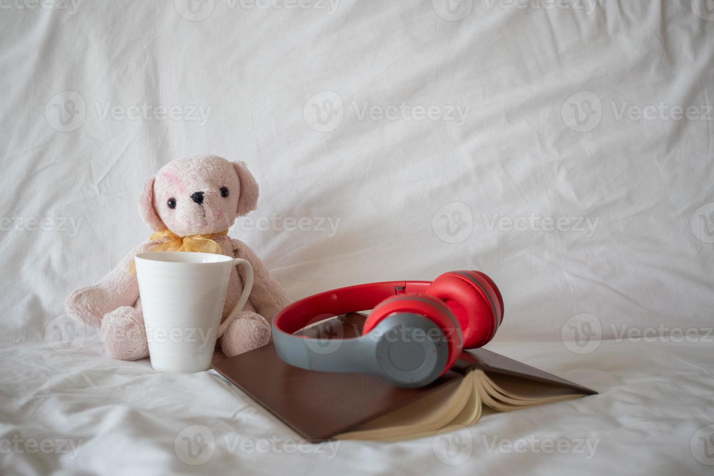 A red headphone is placed on a book near a pink teddy bear on the bed in the morning, set for a comfortable rest. A pair of headphones and a book rests on the white bedding in the bedroom. photo