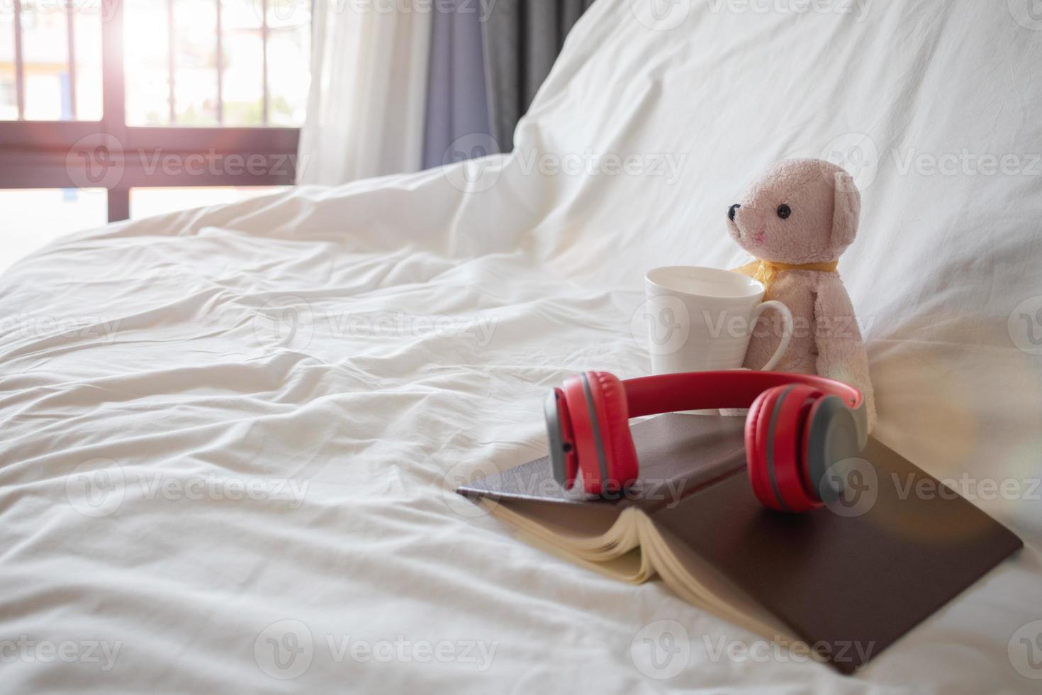 un auricular rojo se coloca en un libro cerca de un oso de peluche rosa en la cama por la mañana, preparado para un descanso cómodo. un par de auriculares y un libro descansan sobre la ropa de cama blanca del dormitorio. foto