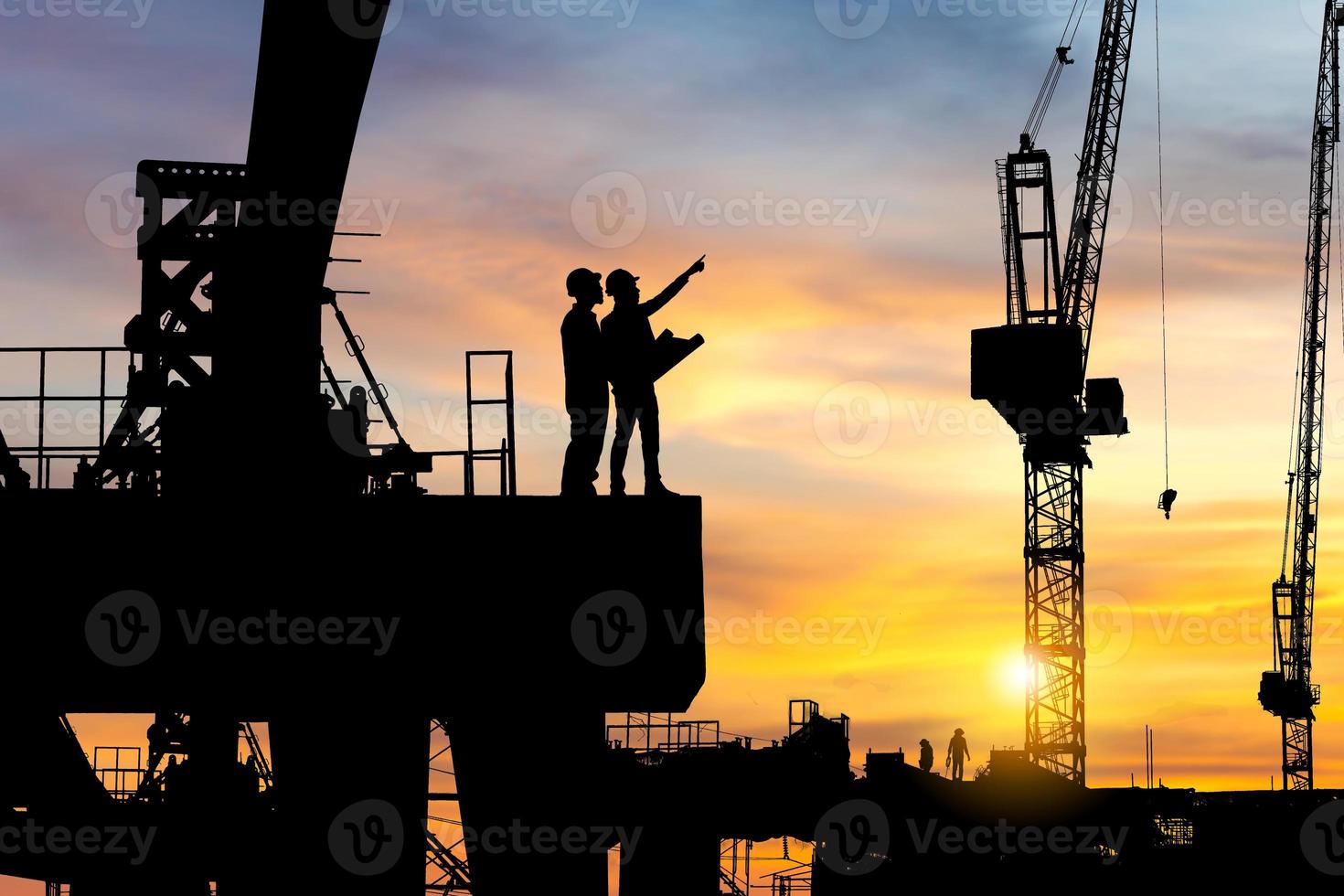 silueta de ingeniero y trabajador revisando el proyecto en el fondo del sitio de construcción de infraestructura pesada, sitio de construcción al atardecer por la noche. foto