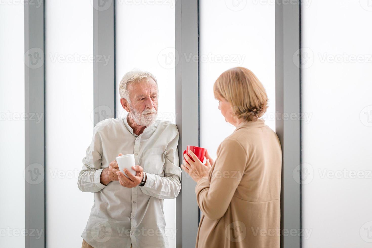 Senior couple inside new home during coffee break, Elderly couple talking while standing near window with cups of coffee photo