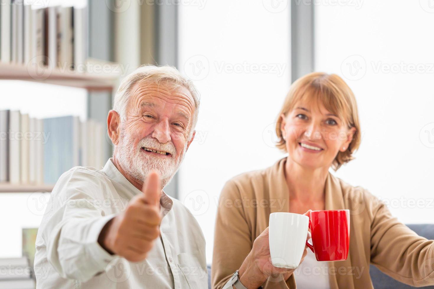 pareja mayor dentro de casa durante un descanso para tomar café, pareja mayor sonriente mostrando los pulgares hacia arriba con tazas de café foto