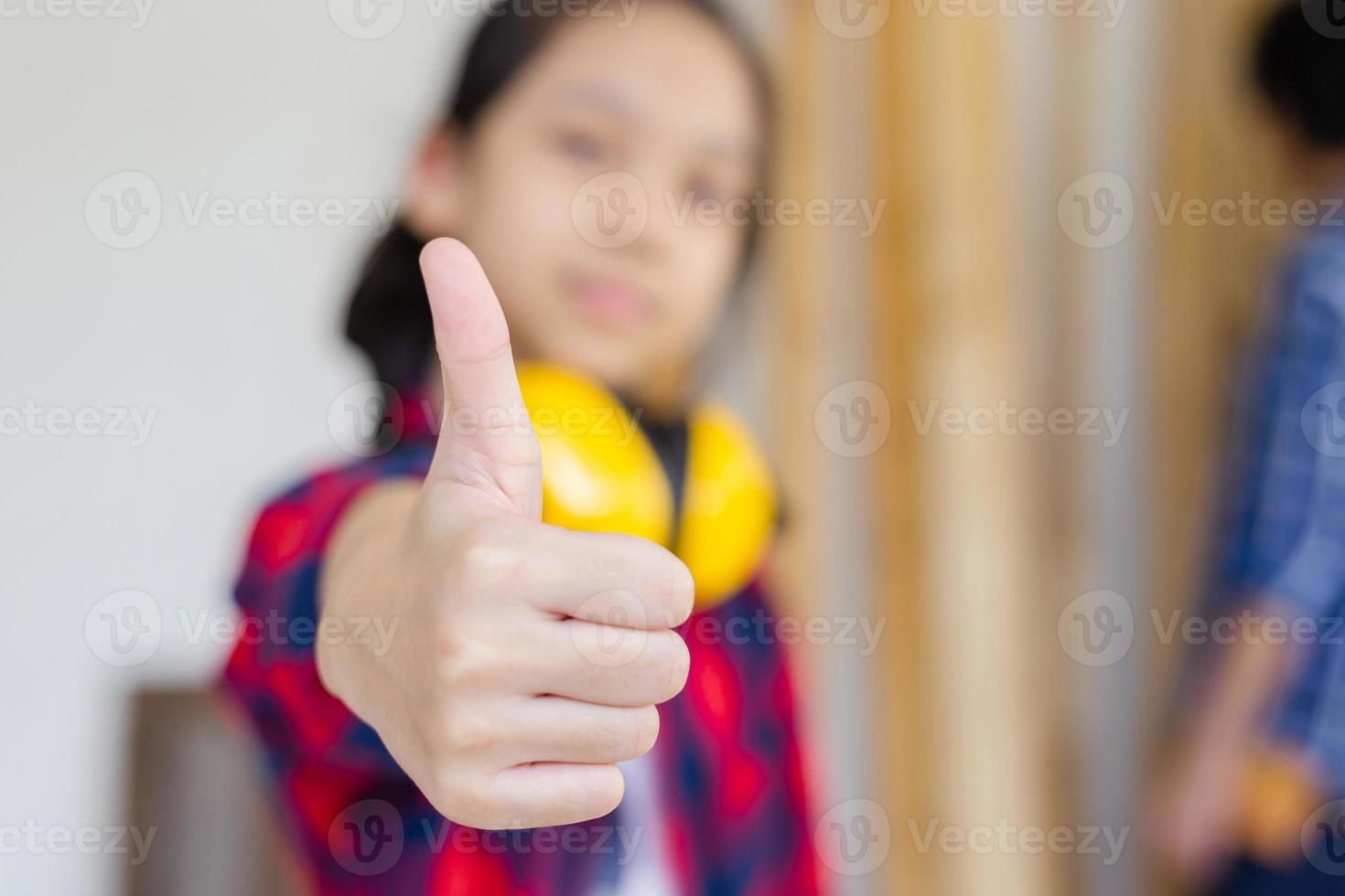 chica asiática de pie dando pulgares hacia arriba como señal de éxito en un taller de carpintería. niño con orejeras de reducción de ruido aprendiendo en el taller artesanal foto