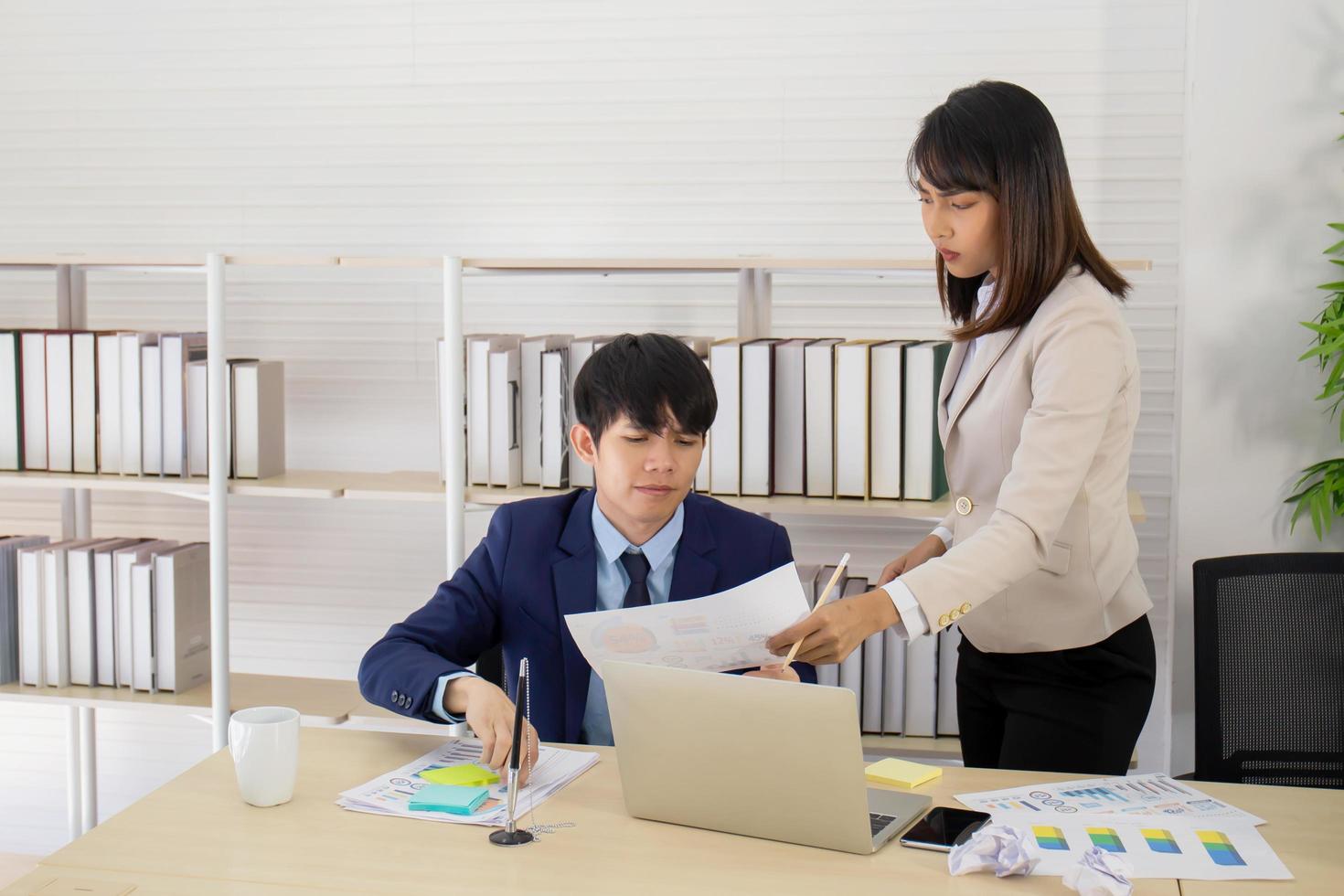 una supervisora asiática se para con un joven empleado en su escritorio con una cara seria. foto