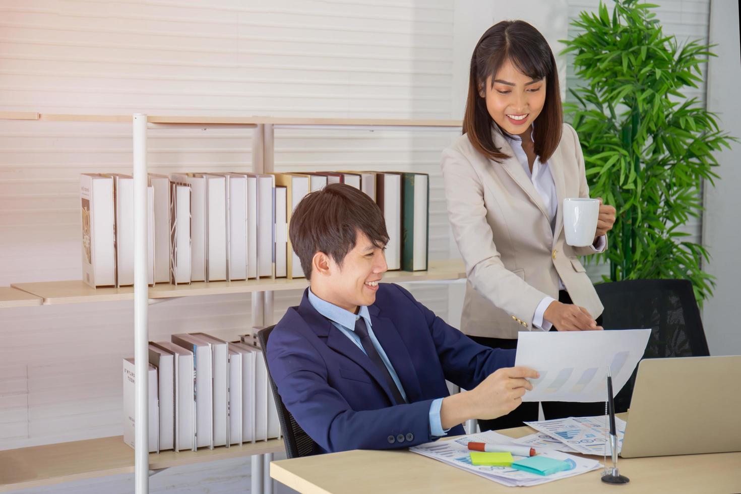 An Asian female coworker stood talking to a business man on the desk in her hand holding a cup of coffee. photo