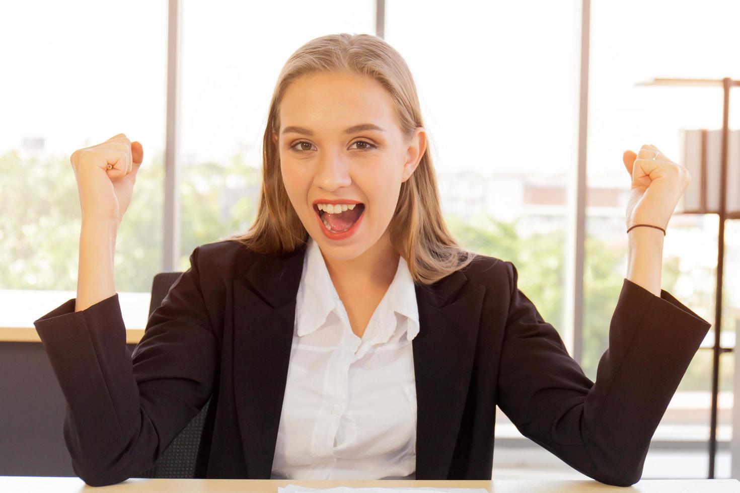 A beautiful business woman in a suit, neatly dressed, sitting in the office, excitedly happy, raised his hand with a bright smile on the table photo