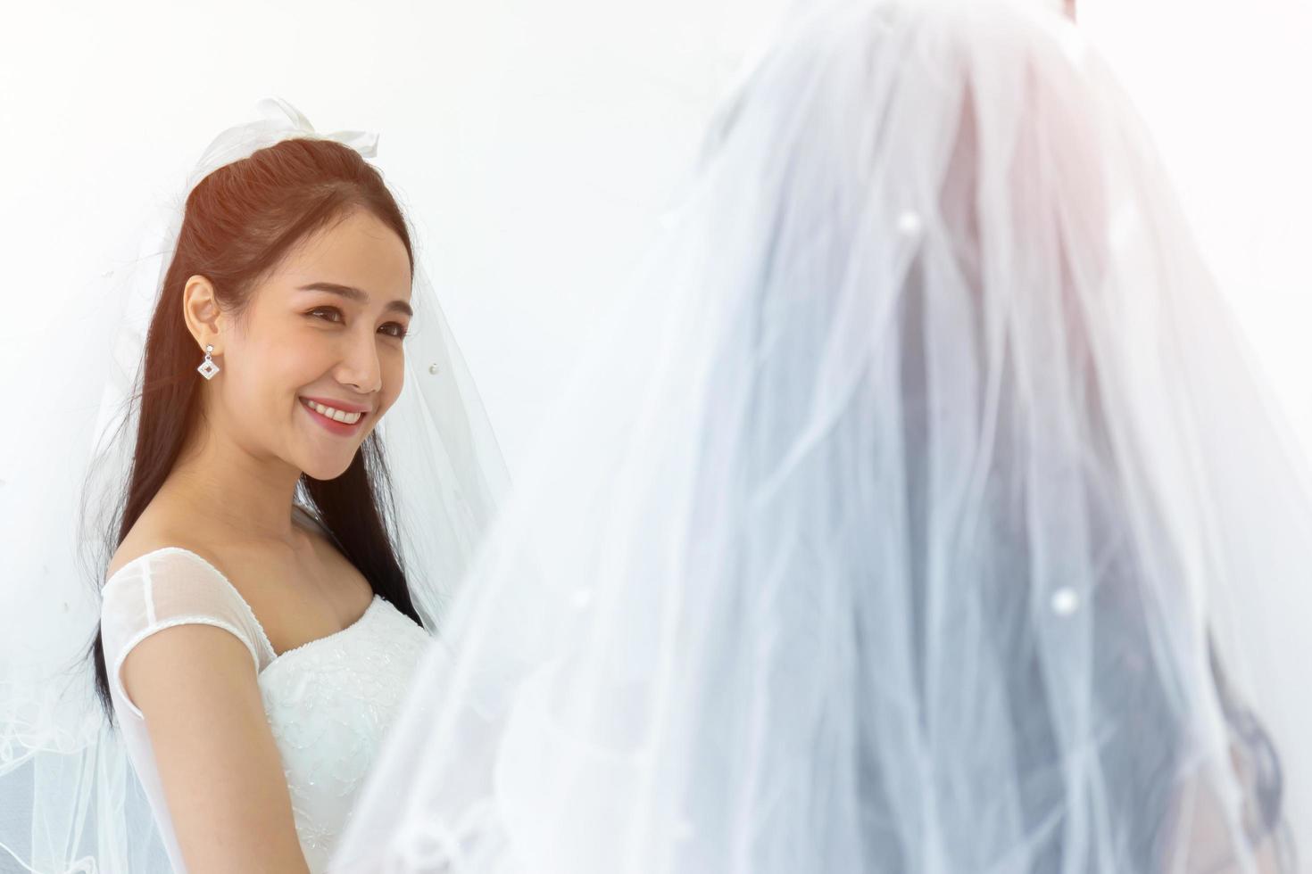 An Asian bride in a white wedding dress stands smiling brightly in front of a mirror reflection. photo