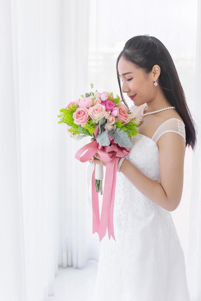 An Asian bride dressed in a white wedding dress stands smiling brightly in a hand holding a beautiful flower bouquet. photo