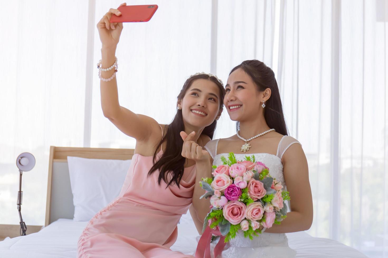 The bride's friend is using a smartphone to take a selfie of an Asian bride holding a bouquet of beautiful flowers in a wedding dress in the dressing room on the bed and making a mini heart hand sign. photo