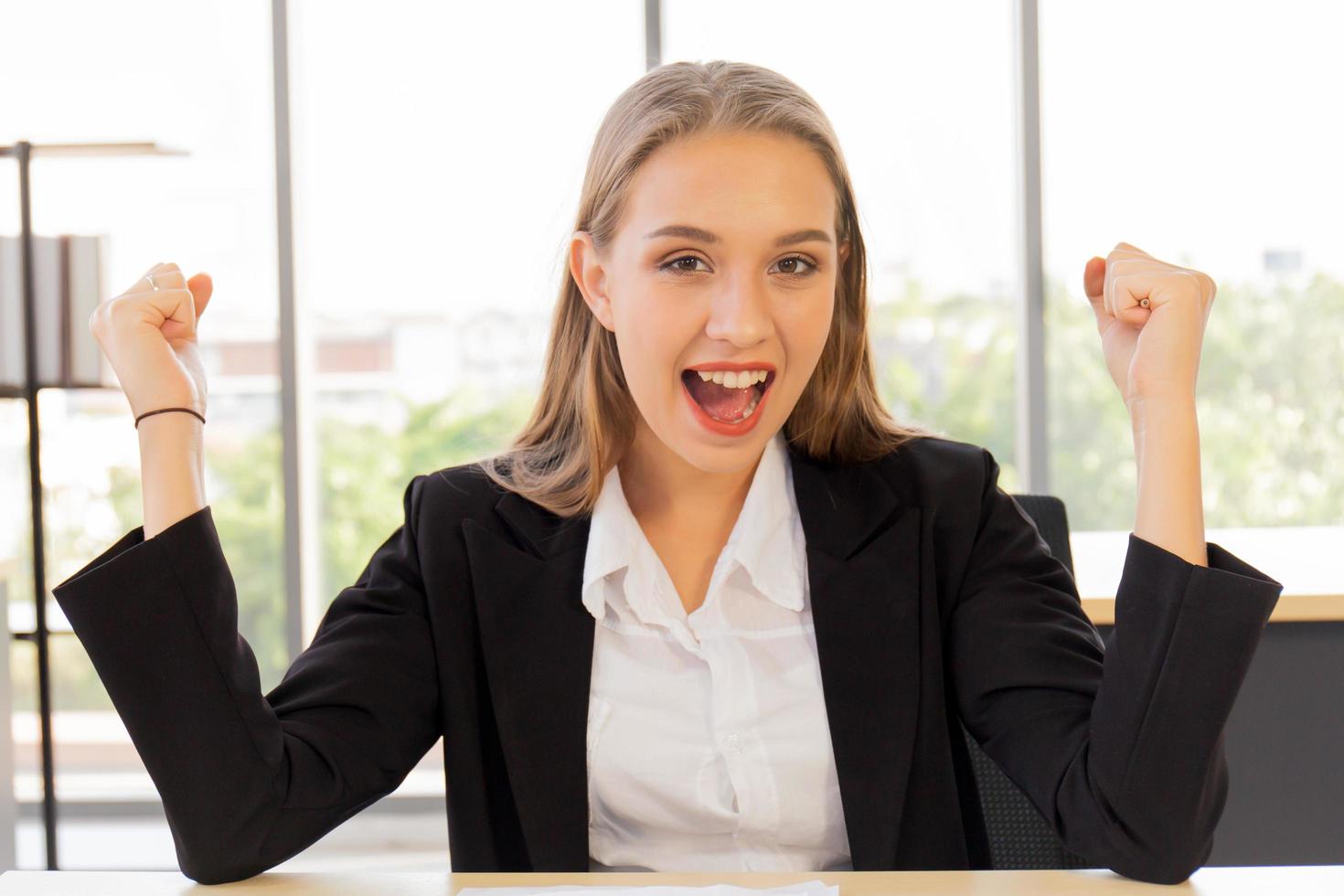 una hermosa mujer de negocios con traje, bien vestida, sentada en la oficina, emocionadamente feliz, levantó la mano con una sonrisa brillante sobre la mesa foto