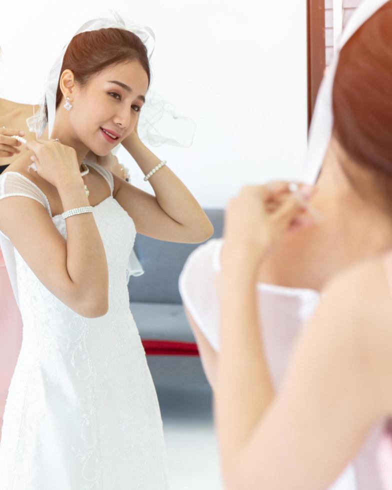 An Asian bride dressed in a white wedding dress is dressed in front of a mirror. photo