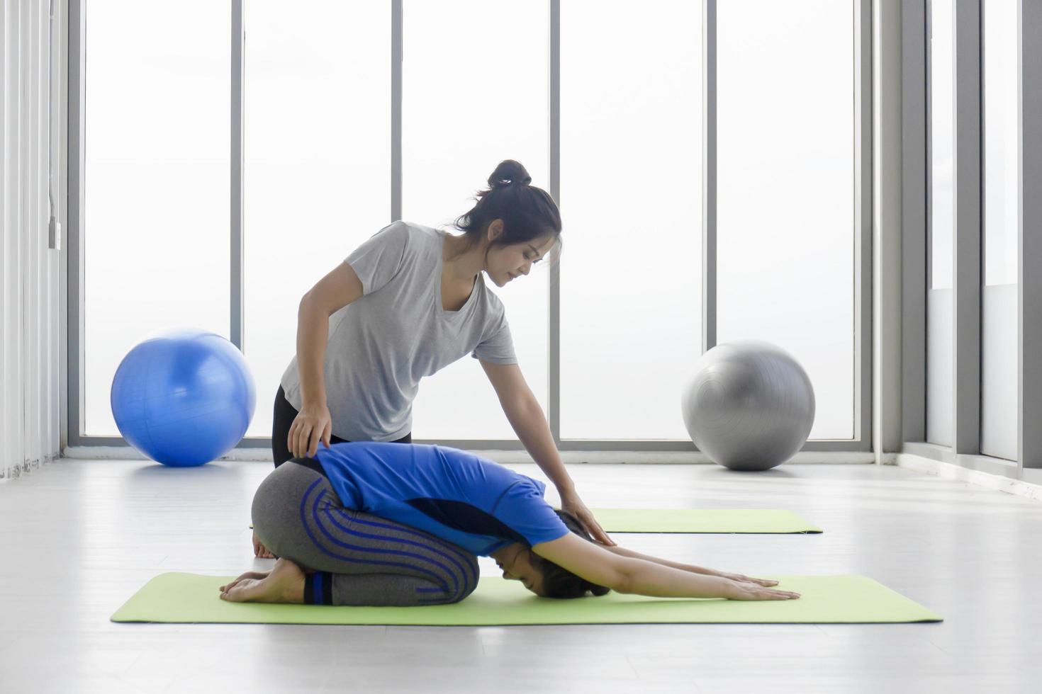 A female trainer is teaching a middle-aged Asian female customer to do yoga on a rubber mat in a gymnasium. photo