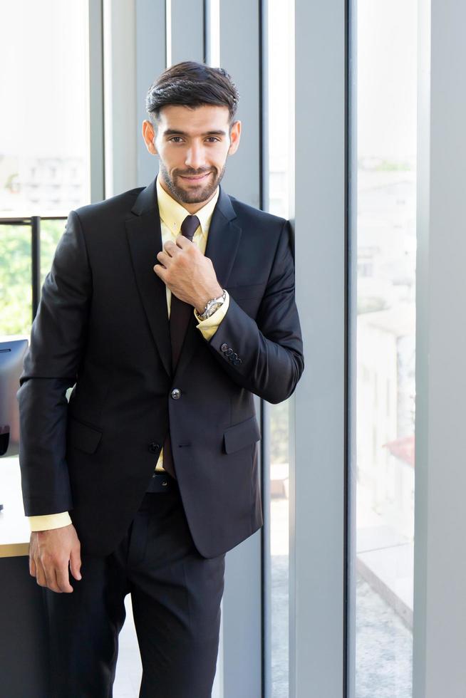 un hombre de negocios con un traje bien vestido se para al lado de la pared de vidrio en la oficina con una sonrisa brillante y una hermosa luz naranja. foto