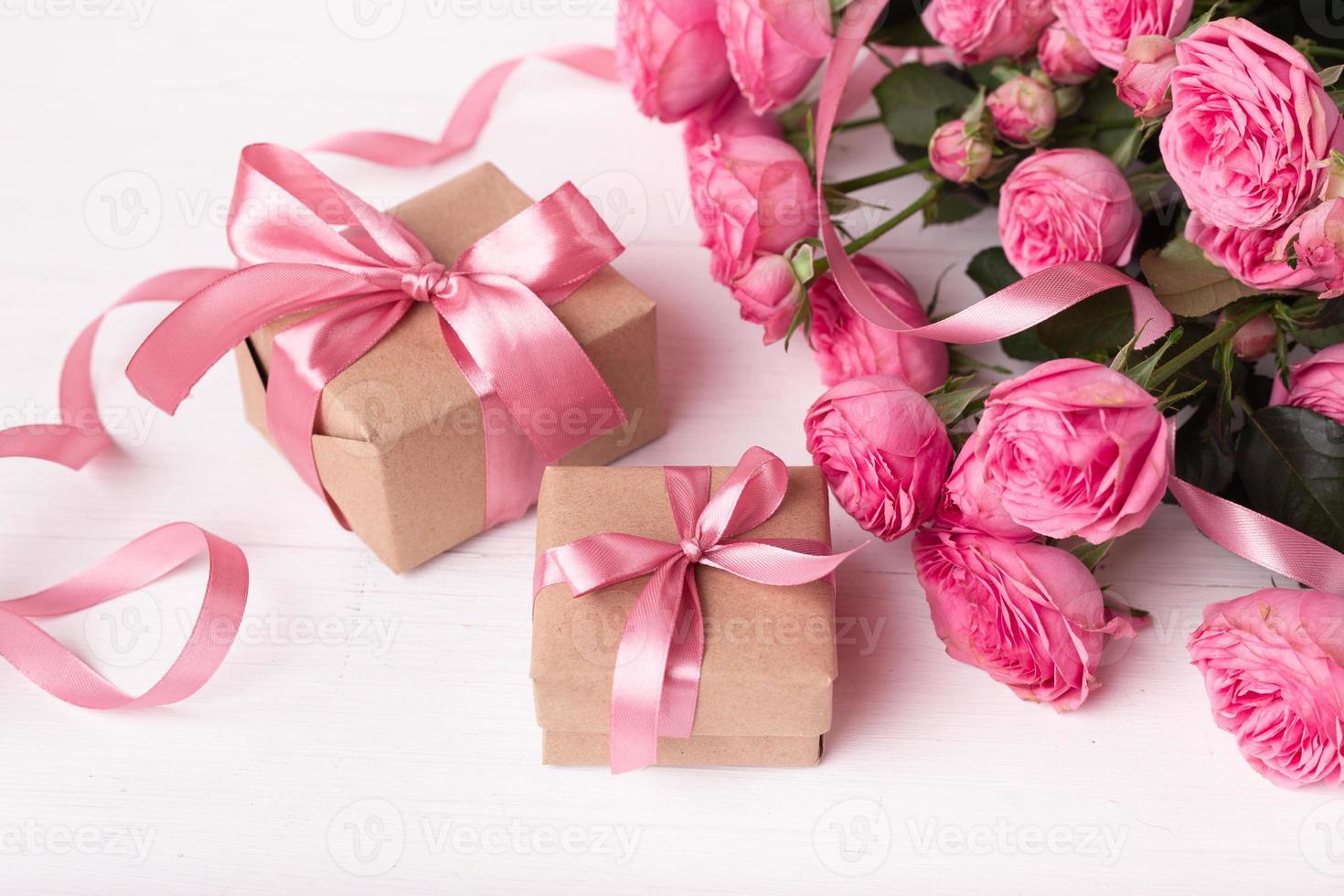 rosas frescas de color rosa pastel y cajas de regalo envueltas en papel kraft con cintas sobre una mesa de madera blanca. foto