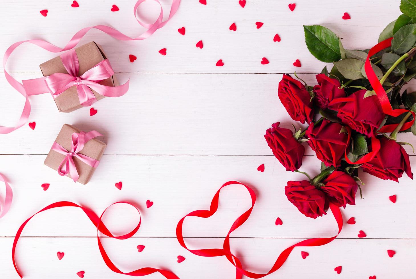 Red ribbon shaped as heart, bouquet of roses, hearts and gift boxes wrapped in kraft papper on white wooden table. photo