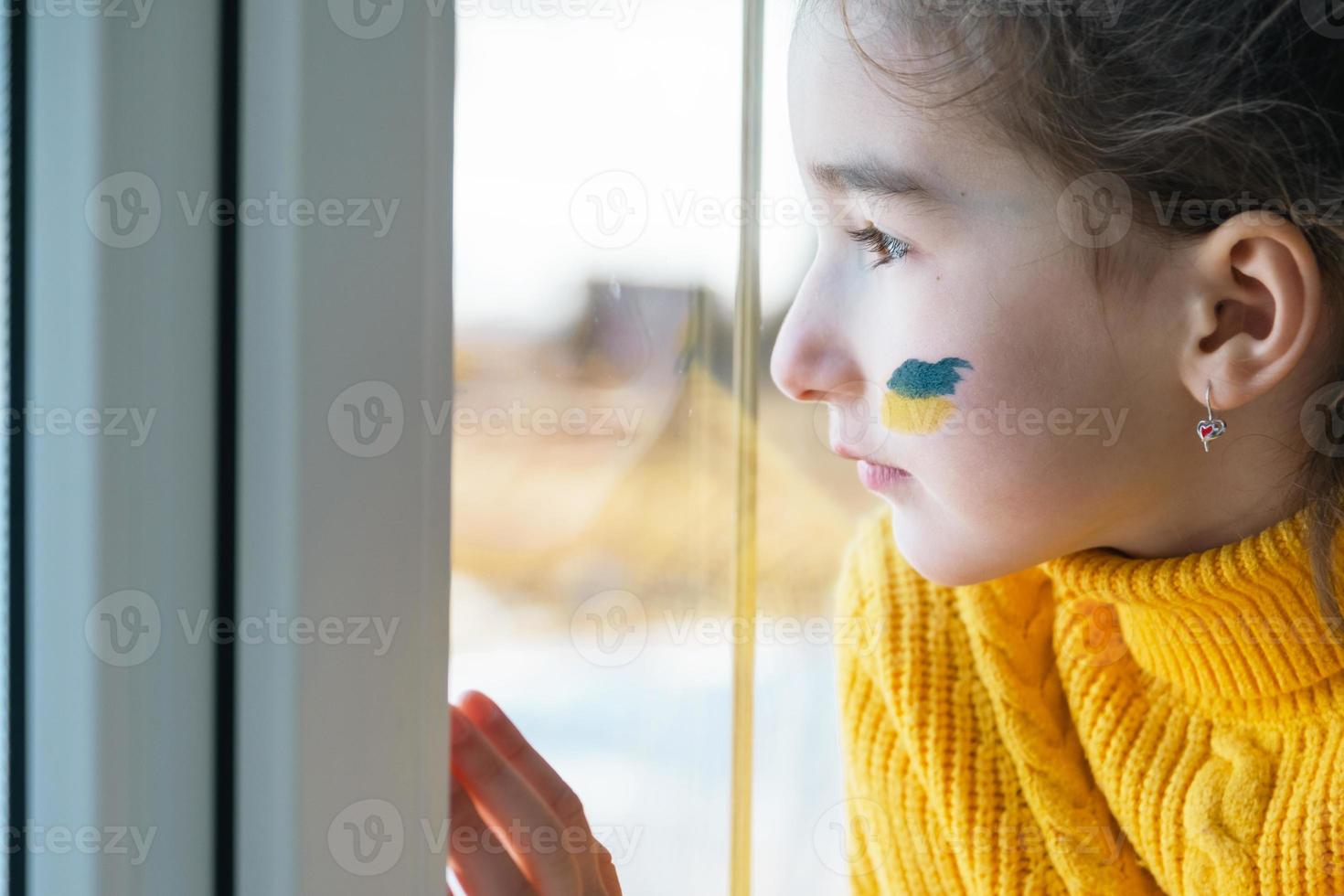 A sad child look at the window with the flag of Ukraine painted on the cheek, worries and fear. Humanitarian aid to children, world peace, security. photo