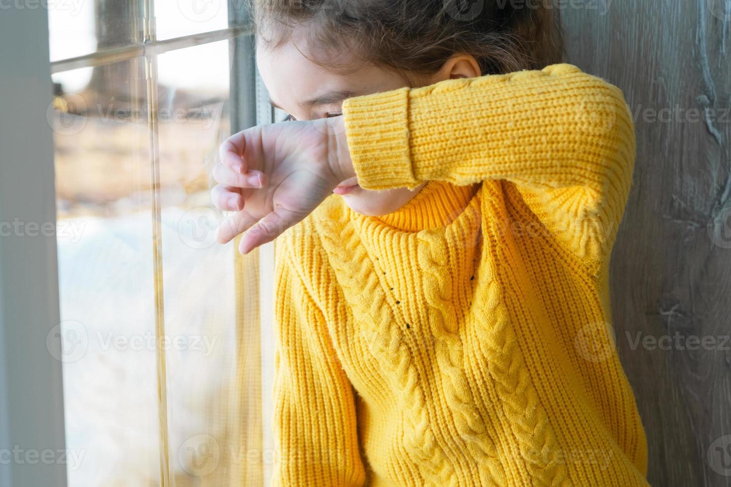 A girl in a yellow sweater looks out the window and covers her face with her hand. Sad child worries, crying and fear photo