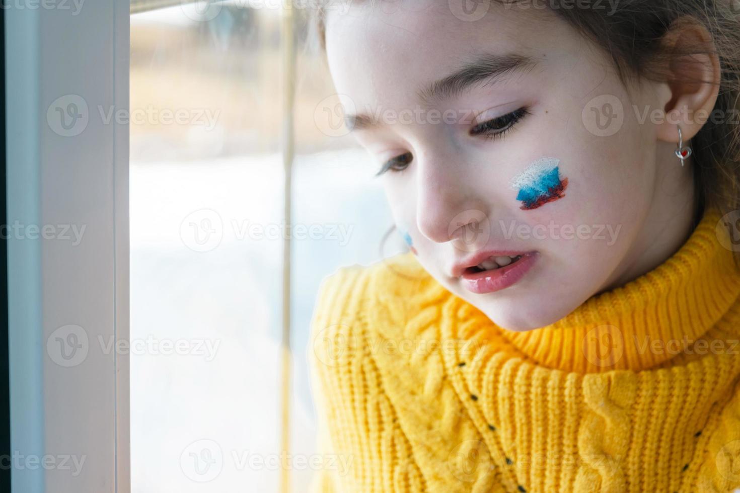 un niño triste en la ventana con la bandera de rusia, se preocupa con lágrimas en los ojos. conflicto entre rusia y ucrania, miedo foto