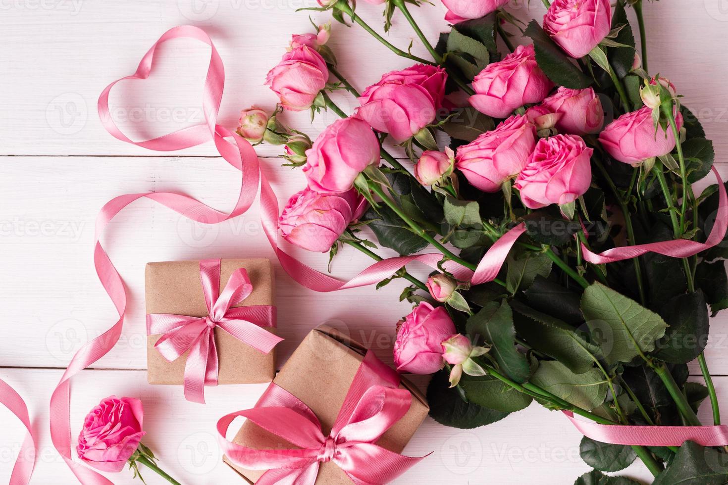 rosas frescas de color rosa pastel y cajas de regalo envueltas en papel kraft con cintas sobre una mesa de madera blanca. foto